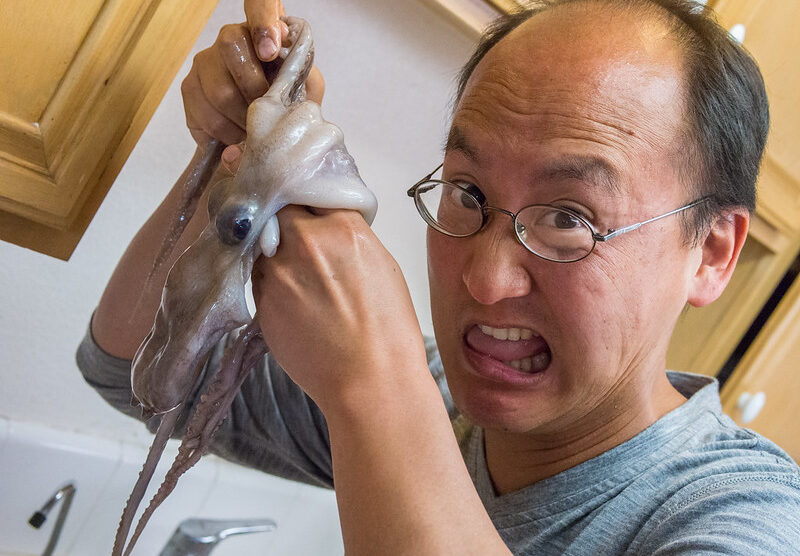 A man with glasses looks surprised and makes a humorous expression while holding a raw octopus in the kitchen. The octopus appears to be squirming in his hand
