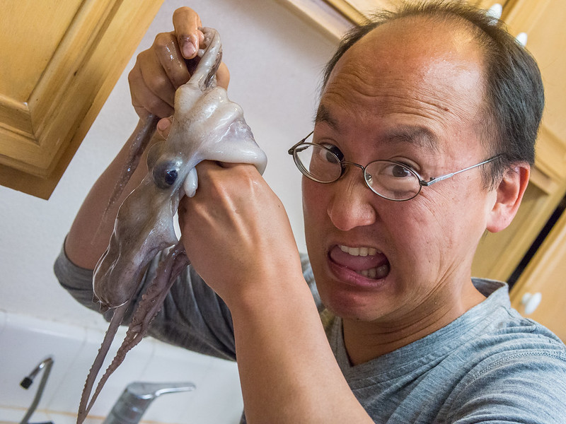A man with glasses looks surprised and makes a humorous expression while holding a raw octopus in the kitchen. The octopus appears to be squirming in his hand