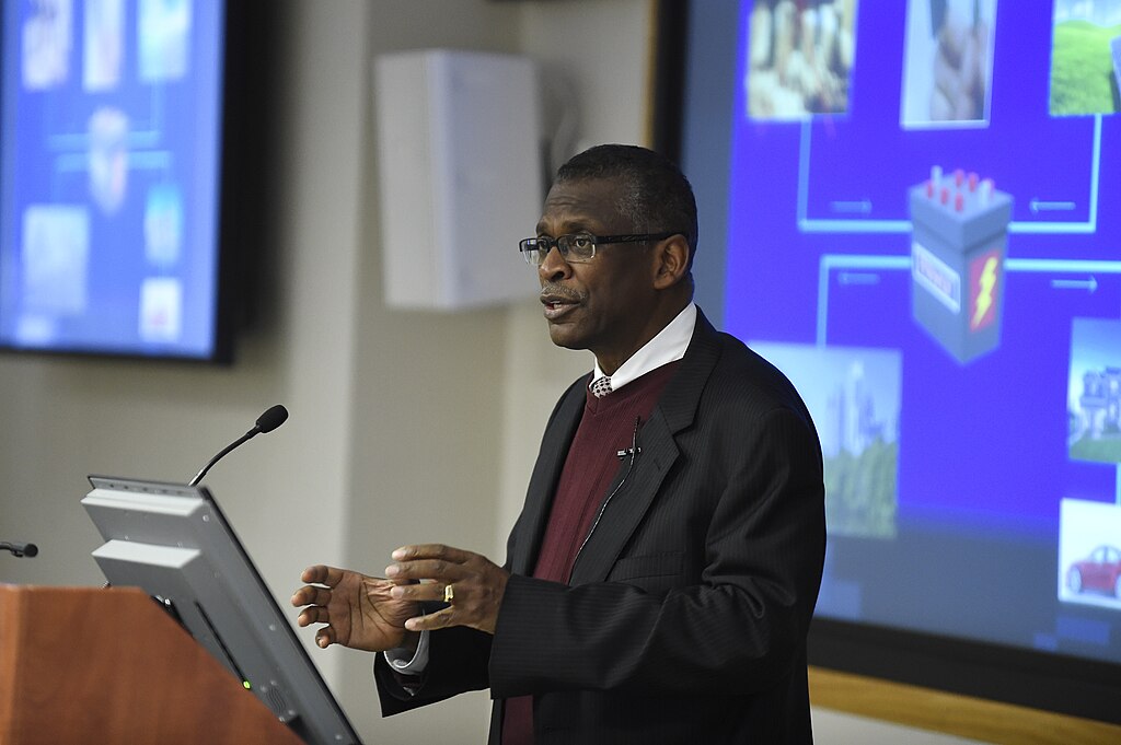 Lonnie Johnson, the inventor and engineer, presenting at a podium with a screen showing a visual presentation in the background.