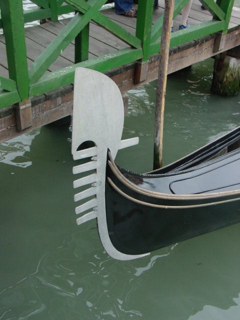Close-up of the metal "fèrro" ornament at the bow of a Venetian gondola near a dock.