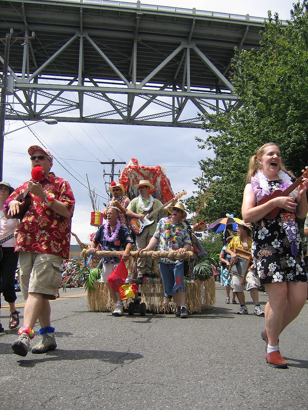 A group of Parrotheads dressed in tropical shirts and leis marches in a parade, some playing instruments like the ukulele. Behind them, a festive float decorated with tropical foliage and flowers features more people playing guitars and singing. The group parades under a large bridge, with bystanders watching from the sidelines.