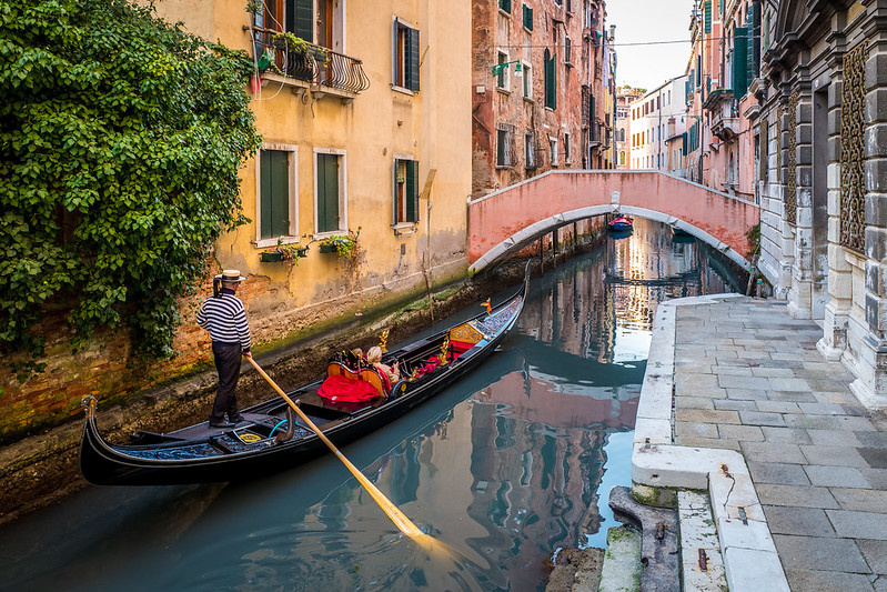 Gondola with a gondolier and tourists on a calm Venetian canal, surrounded by traditional buildings and a small arched bridge.