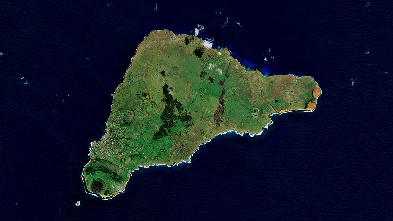Satellite image of Easter Island, showing its green volcanic landscape surrounded by dark blue ocean, with visible coastal cliffs and varying terrain.