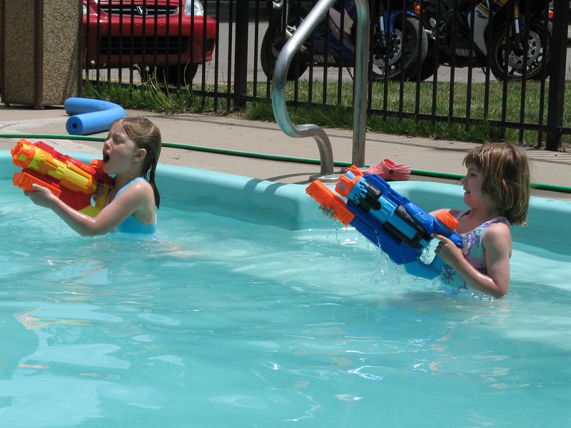  Two young children playing in a pool with large Super Soaker water guns, engaged in a water battle on a sunny day.