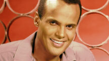 A close-up portrait of a smiling man with short hair, wearing a light-colored shirt. The background features circular patterns in a warm hue.