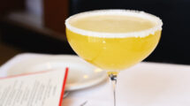 A classic Margarita cocktail served in a coupe glass with a salted rim, resting on a white tablecloth next to a menu and a plate.