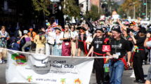 A colorful parade scene featuring members of the Left Coast Parrot Head Club, "partying with a purpose." Many members are dressed in tropical-themed attire and pirate costumes. The group is holding a banner with their club name, promoting their activities in Ventura County and beyond. The street is filled with people, some in beach-inspired clothing, celebrating in a festive, community-driven atmosphere.