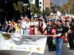A colorful parade scene featuring members of the Left Coast Parrot Head Club, "partying with a purpose." Many members are dressed in tropical-themed attire and pirate costumes. The group is holding a banner with their club name, promoting their activities in Ventura County and beyond. The street is filled with people, some in beach-inspired clothing, celebrating in a festive, community-driven atmosphere.