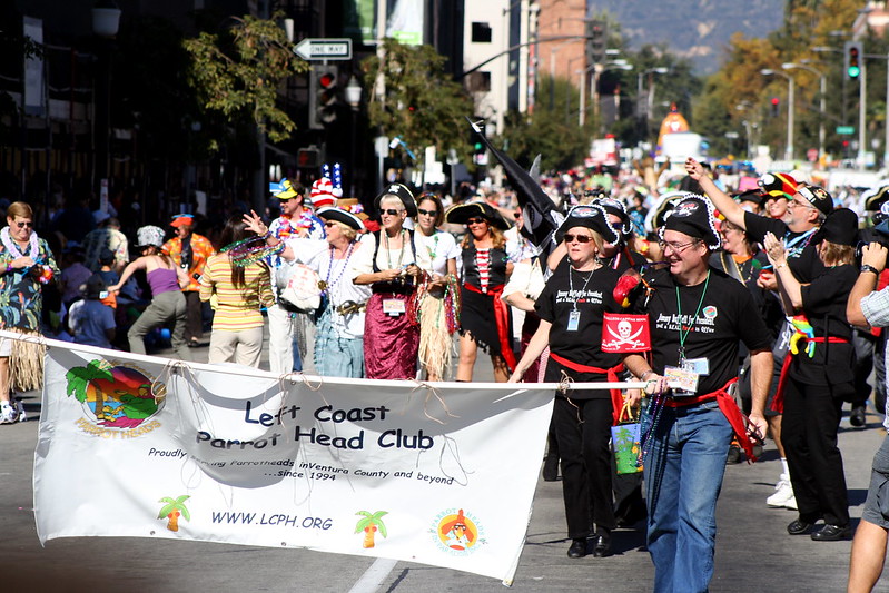 A colorful parade scene featuring members of the Left Coast Parrot Head Club, "partying with a purpose." Many members are dressed in tropical-themed attire and pirate costumes. The group is holding a banner with their club name, promoting their activities in Ventura County and beyond. The street is filled with people, some in beach-inspired clothing, celebrating in a festive, community-driven atmosphere.