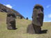 Close-up of two Moai statues on a grassy slope under a blue sky, partially buried in the ground at Rano Raraku quarry on Easter Island.