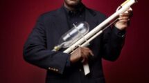 An inventor holding an early prototype of a Super Soaker water gun against a dark red background.