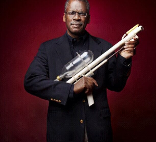 An inventor holding an early prototype of a Super Soaker water gun against a dark red background.