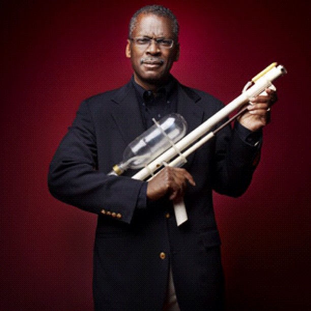 An inventor holding an early prototype of a Super Soaker water gun against a dark red background.
