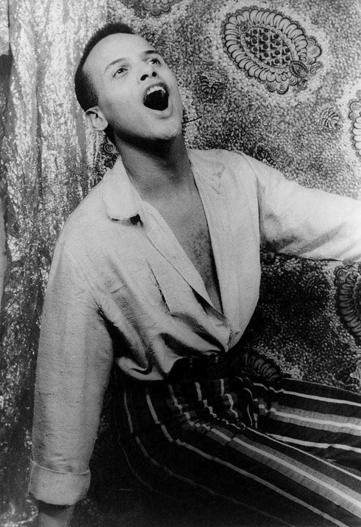  Black and white photo of Harry Belafonte singing passionately with his mouth open, wearing a loose-fitting shirt and striped pants, against a patterned backdrop.