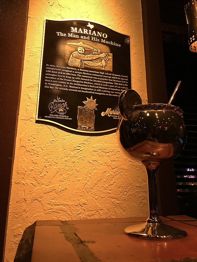 A plaque titled "Mariano: The Man and His Machine" detailing the invention of the frozen margarita machine by Mariano Martinez, mounted on a textured yellow wall beside a margarita glass.