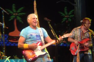 A smiling Jimmy Buffett wearing a tropical shirt and playing an electric guitar stands on stage with a microphone. Palm trees and a tropical background are visible behind him. Another man is playing a bass guitar in the background.