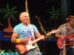 A smiling Jimmy Buffett wearing a tropical shirt and playing an electric guitar stands on stage with a microphone. Palm trees and a tropical background are visible behind him. Another man is playing a bass guitar in the background.