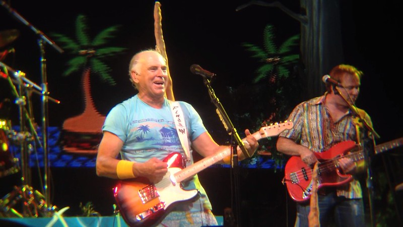 A smiling Jimmy Buffett wearing a tropical shirt and playing an electric guitar stands on stage with a microphone. Palm trees and a tropical background are visible behind him. Another man is playing a bass guitar in the background.