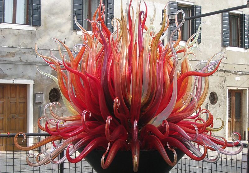A vibrant glass sculpture in Murano, Italy, with red, orange, and yellow glass spikes resembling flames in front of a traditional Venetian building.