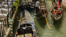 Gondolas on a narrow canal in Venice, with gondoliers navigating and tourists seated, surrounded by buildings and a small bridge.