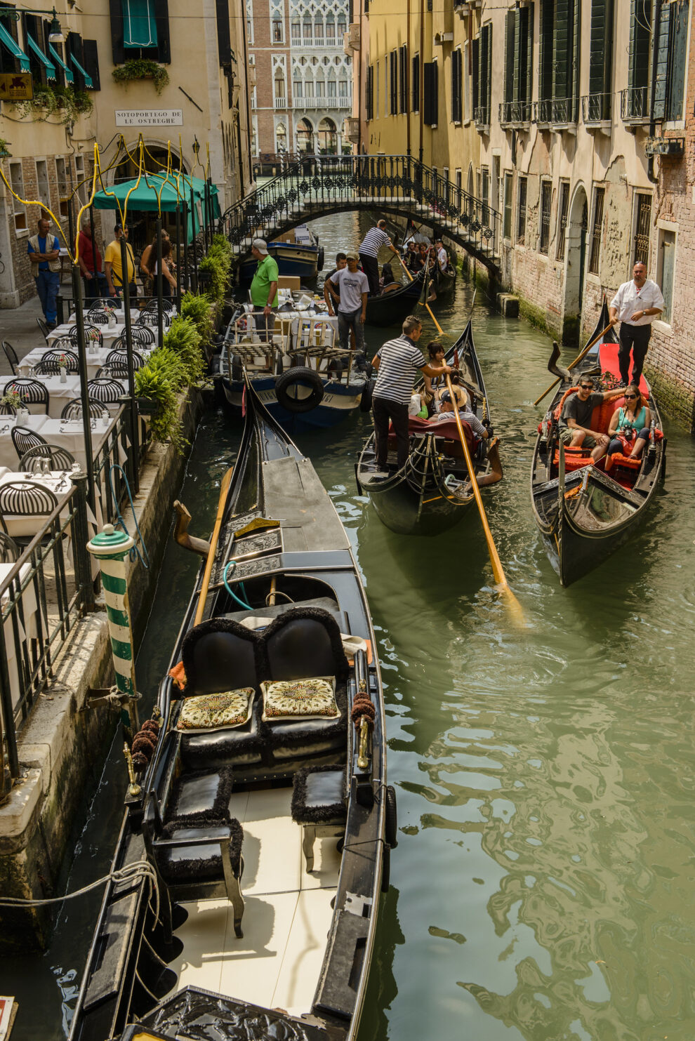 Gondolas on a narrow canal in Venice, with gondoliers navigating and tourists seated, surrounded by buildings and a small bridge.