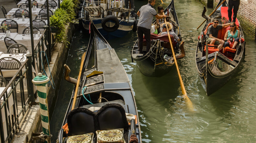 Gondolas on a narrow canal in Venice, with gondoliers navigating and tourists seated, surrounded by buildings and a small bridge.