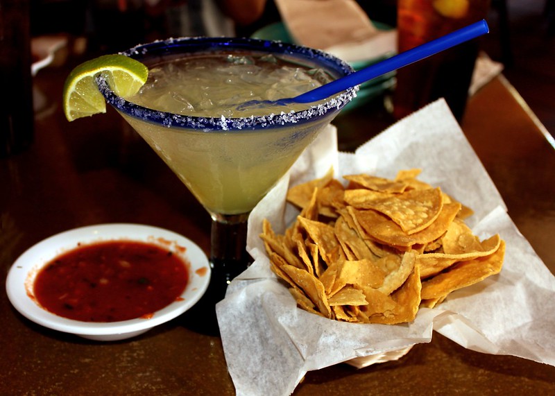A Margarita served in a large glass with a salted rim and a lime wedge, accompanied by a basket of tortilla chips and a small dish of salsa.