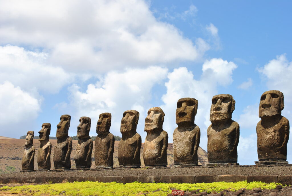 : A row of 15 large Moai statues standing on a stone platform at Ahu Tongariki on Easter Island, with a bright blue sky and clouds in the background. The statues, carved from volcanic rock, represent the Rapa Nui people's ancestors.