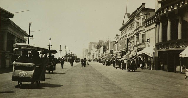 A vintage photograph of the Atlantic City Boardwalk, with people strolling and rolling chairs visible from the 1920s.