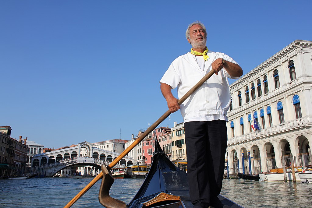 Gondolier rowing on the Grand Canal in Venice, with the Rialto Bridge and historic buildings in the background.