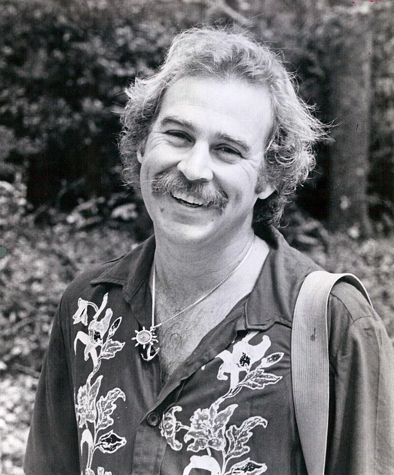 A black and white portrait of a man with a mustache, smiling warmly at the camera. He is wearing a floral shirt and a sun-shaped necklace, with trees and foliage in the background.