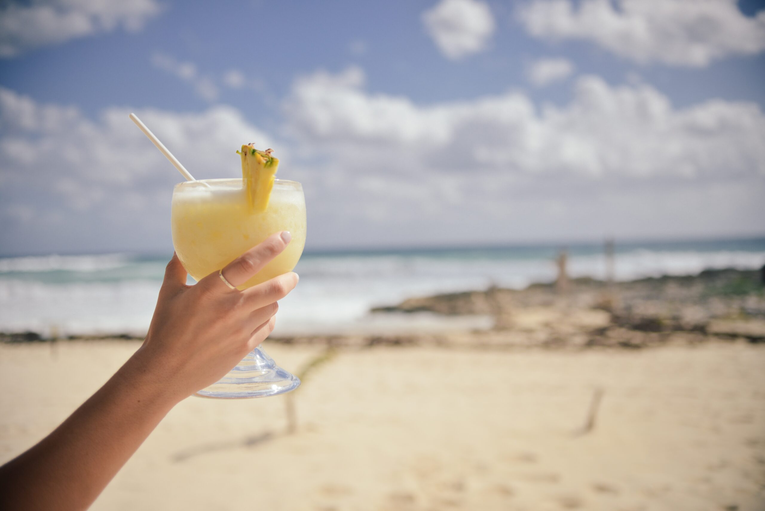 Hand holding a Piña Colada topped with a pineapple wedge on a sunny beach, with the ocean in the background.