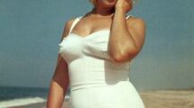Marilyn Monroe on a beach in a white swimsuit, posing gracefully with her hand near her face, against an ocean backdrop