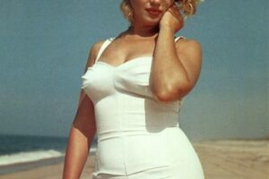 Marilyn Monroe on a beach in a white swimsuit, posing gracefully with her hand near her face, against an ocean backdrop