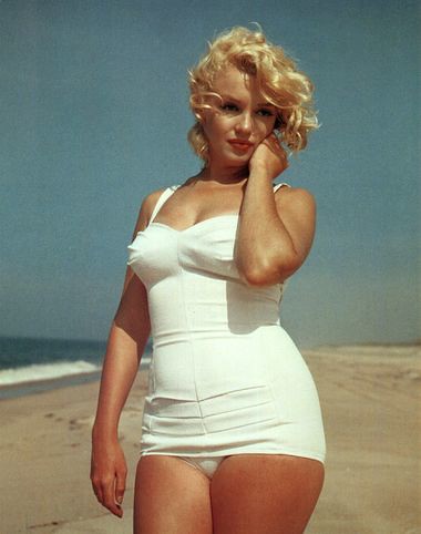 Marilyn Monroe on a beach in a white swimsuit, posing gracefully with her hand near her face, against an ocean backdrop