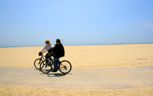 A couple riding bicycles on a paved path across a sandy beach under a clear blue sky, encouraging exploration of potential dream destinations