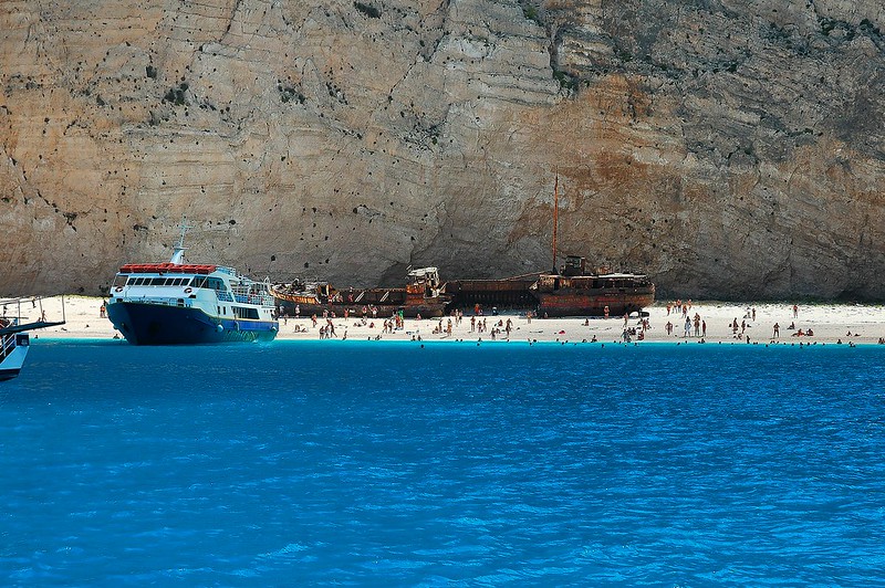 A shipwreck on the shore of Navagio Beach, surrounded by steep cliffs and clear blue water.