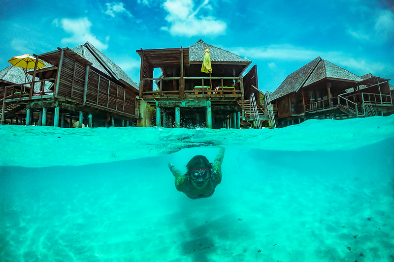 A person snorkeling in the clear turquoise water with overwater bungalows in the Maldives in the background.