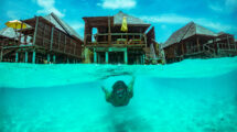 A person snorkeling underwater with overwater bungalows in the Maldives in the background.