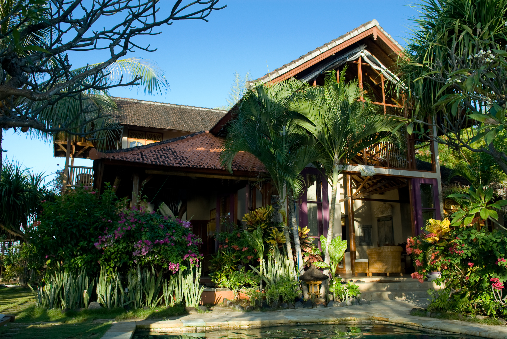 A tropical villa with a terracotta roof surrounded by palm trees, vibrant flowers, and greenery, with a small pool in the foreground.