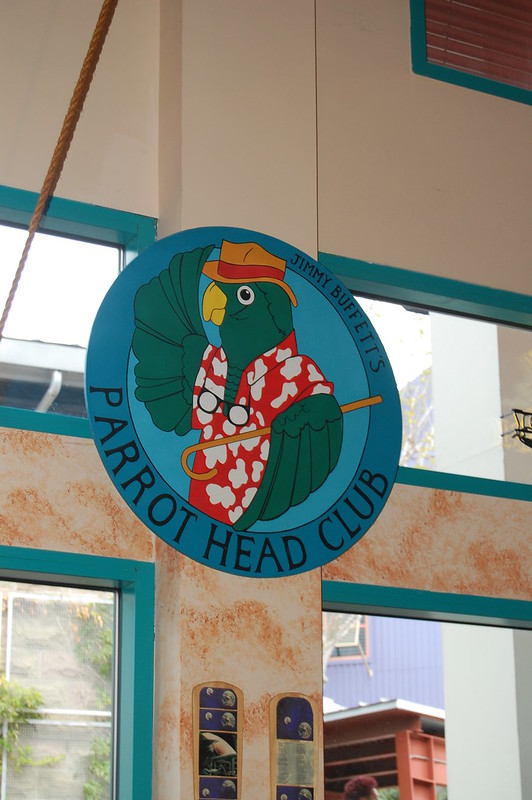 A sign for Jimmy Buffett's Parrot Head Club, showing a green parrot wearing a red Hawaiian shirt, straw hat, and sunglasses.