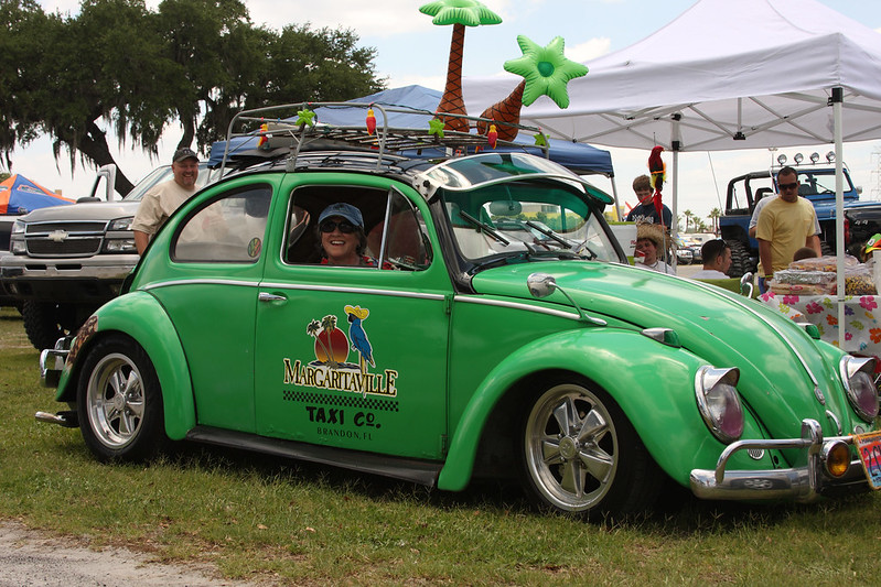 A green Volkswagen Beetle with a Margaritaville theme, decorated with inflatable palm trees, and the words 'Margaritaville Taxi Co.' on the side, parked at a car show.