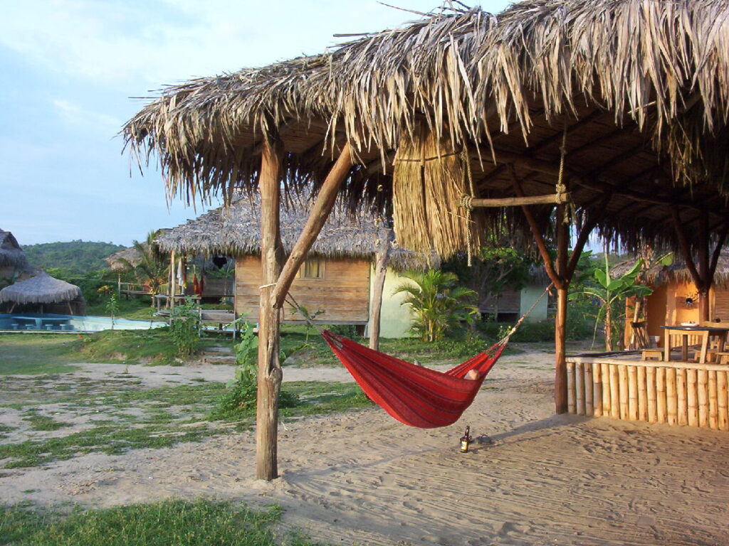 A red hammock hangs under a thatched-roof hut in a tropical setting with sandy ground, lush greenery, and rustic bamboo structures, evoking a peaceful paradise.