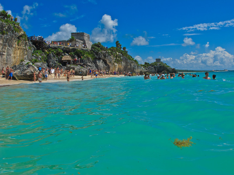 Clear turquoise waters of Tulum Beach with ancient Mayan ruins on the cliff in the background and tourists swimming.