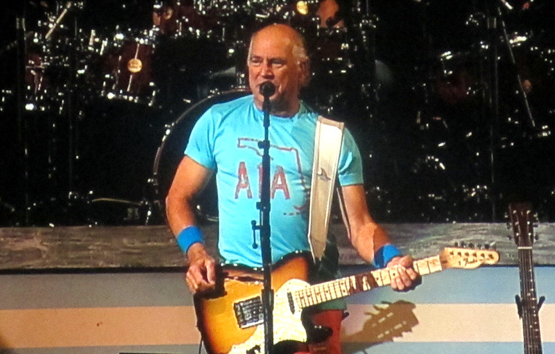 Jimmy Buffett wearing a light blue shirt, playing guitar on stage, with drums visible in the background.