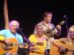 Jimmy Buffett, wearing a blue shirt, performs with his Coral Reefers Band. He plays guitar alongside musicians playing ukulele, percussion, and another guitar.