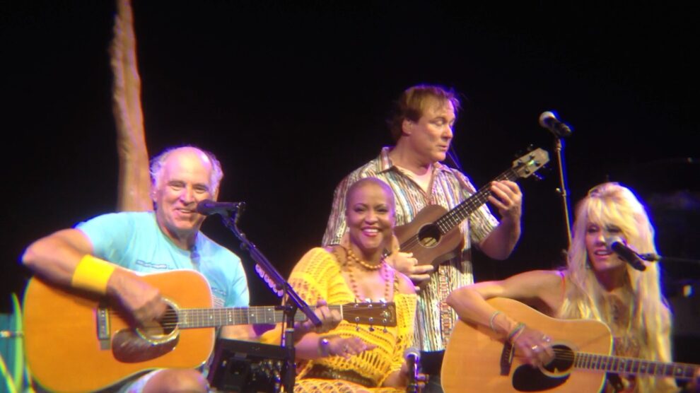 Jimmy Buffett, wearing a blue shirt, performs with his Coral Reefers Band. He plays guitar alongside musicians playing ukulele, percussion, and another guitar.