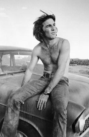 Dennis Wilson shirtless, sitting on a vintage car's hood in a black-and-white photo, with a rural background.