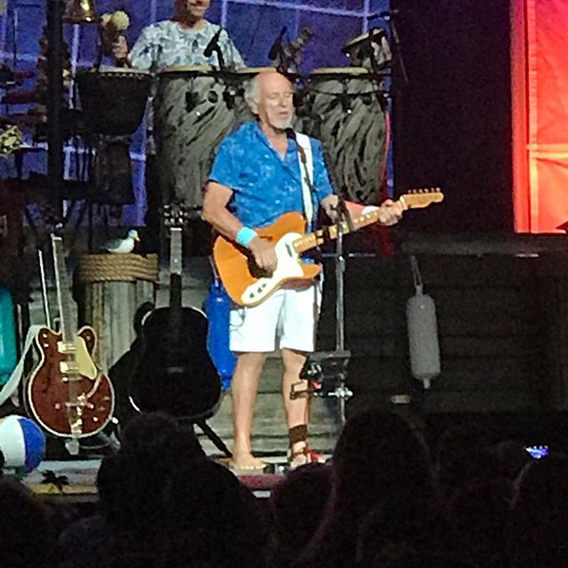 Jimmy Buffett performing on stage in a blue Hawaiian shirt and white shorts, playing an orange electric guitar with tropical props and drums in the background.