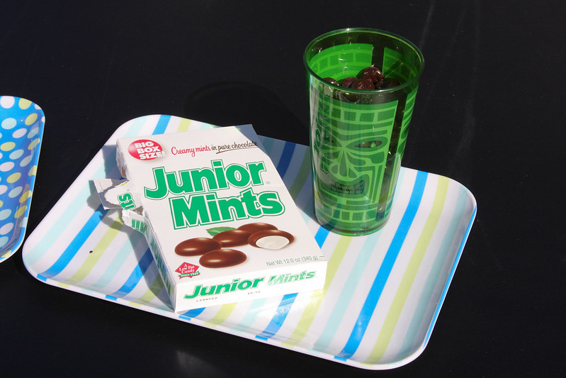 A box of Junior Mints alongside a green tiki-themed cup filled with chocolates, displayed on a striped tray with bright colors.